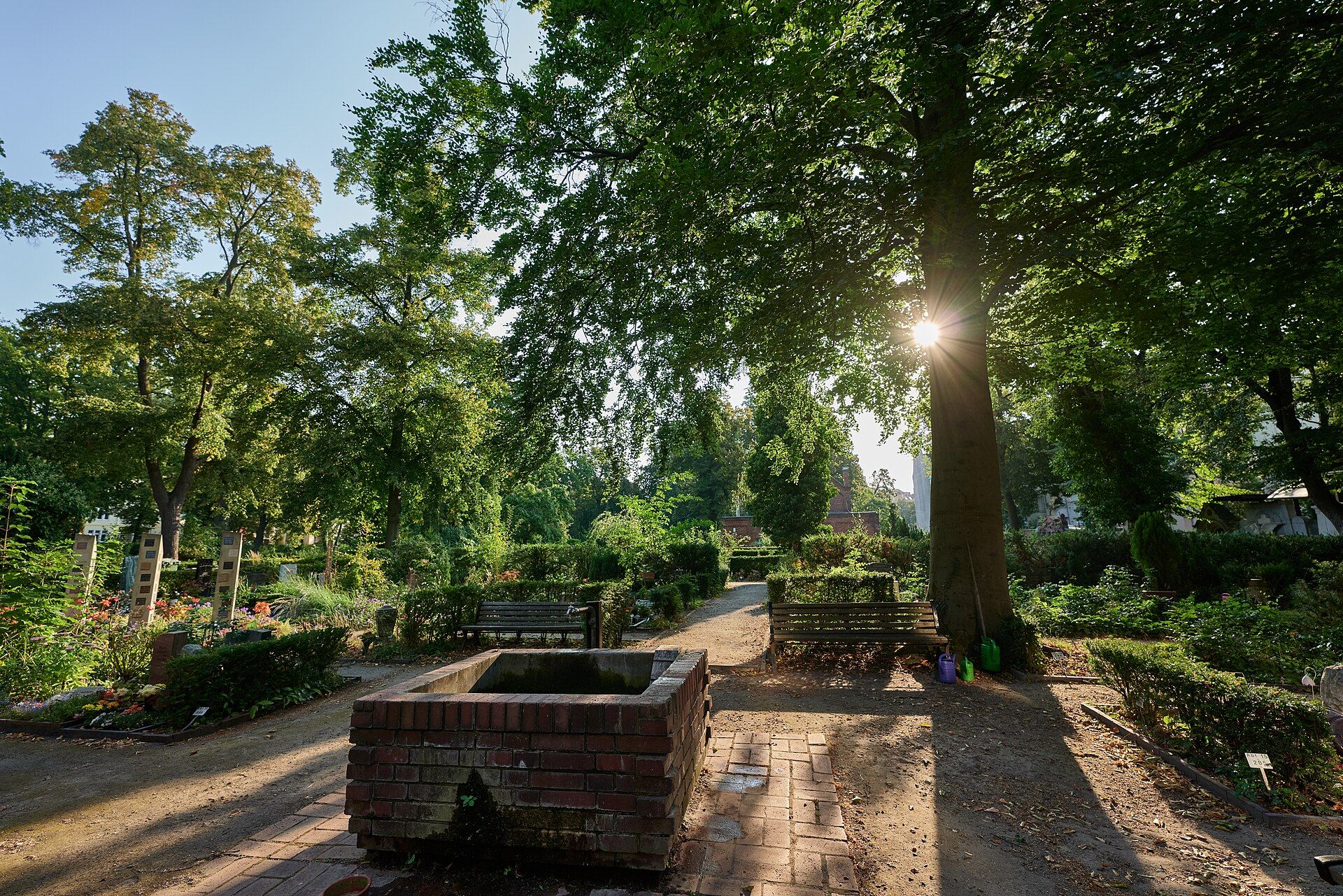 Gemauerte Wasserstelle und Bänke zwischen bepflanzten Gräbern auf dem Friedhof Stubenrauchstraße